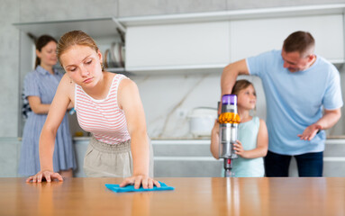 Joint family house cleaning, household tasks, household help. Eldest girl wipe dry kitchen table, father with little daughter vacuums floor in background, mother mop dry dishes