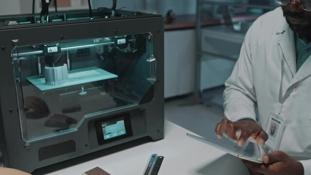Zoom out shot of engineer in white coat sitting in laboratory testing 3D printing machine with help of digital tablet