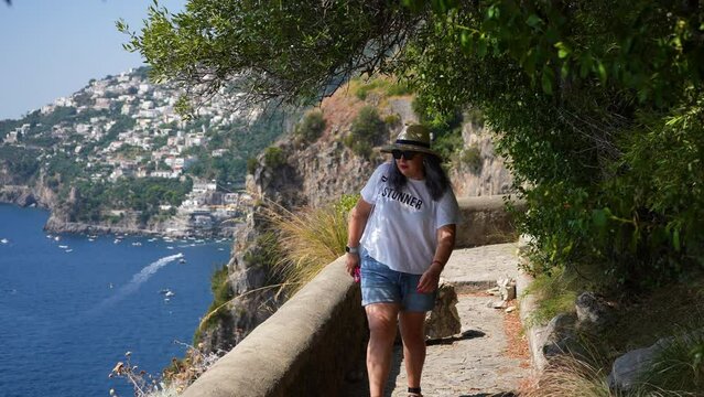 Happy female tourist with backpack in sunglasses and hat walking through path next to coast and sea, stops and taking picture, on background of beautiful Amalfi urban and seascape on summer day,