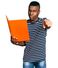 Young african american man holding book pointing with finger to the camera and to you, confident gesture looking serious