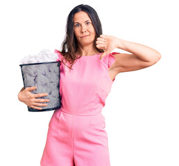 Young beautiful brunette woman holding paper bin full of crumpled papers with angry face, negative...