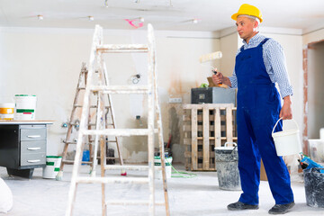 Man painter in coverall and hardhat carrying bucket and paint roller. Male builder working in...