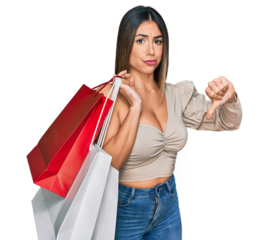 Young hispanic woman holding shopping bags with angry face, negative sign showing dislike with thumbs down, rejection concept