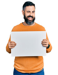 Hispanic man with beard holding blank empty banner winking looking at the camera with sexy expression, cheerful and happy face.