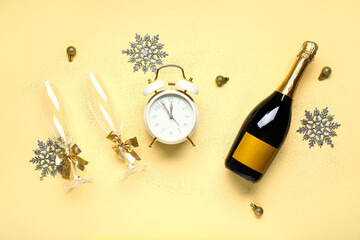 Alarm clock with Christmas balls, glasses and bottle of champagne on yellow background