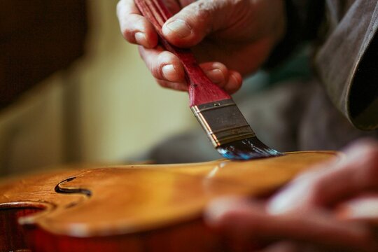 Senior expert violin maker luthier wrinkled hand hold brush, varnish classic handmade violin paint natural ingredient recipe in Cremona Italy home of best artisan of this kind, selective focus, bokeh