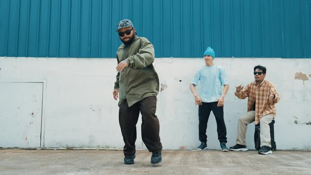 Skilled hipster doing cart wheel while multicultural friends encourage him. Hip-hop dancer moving foot step to music surrounded by group of hipster at wall near street. Outdoor sport 2024. hiphop.