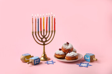 Menorah, dreidels and plate with donuts for Hanukkah celebration on pink background