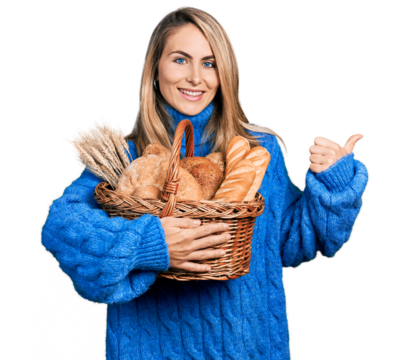 Young blonde woman holding wicker basket with bread pointing thumb up to the side smiling happy with open mouth