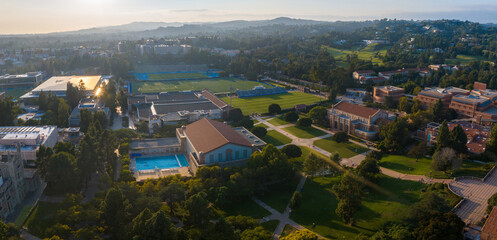 Aerial view of UCLA campus bathed in golden light, featuring Gothic architecture, a blue-roofed pool, green hills, and a clear, sunny sky, amidst a vibrant, verdant landscape.