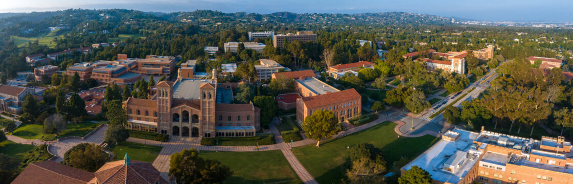 Aerial view of UCLA campus in Westwood, Los Angeles, showcasing Romanesque Revival architecture, modern buildings, green spaces, and a serene city backdrop under a partly cloudy sky.