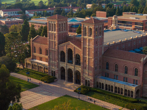 Aerial view of UCLA campus with Royce Hall center stage, Romanesque architecture, green spaces, and Westwood's urban backdrop in golden morning - afternoon light.