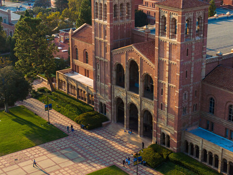 Aerial view of UCLA's Royce Hall, featuring Gothic Revival architecture with twin towers, arched windows, and a red-tiled roof, bathed in golden sunlight on a clear day.