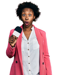 African american woman with afro hair holding reporter microphone scared and amazed with open mouth...