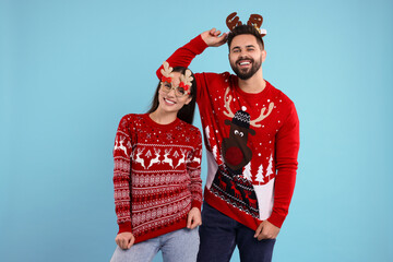 Happy young couple in Christmas sweaters, reindeer headband and funny glasses on light blue...