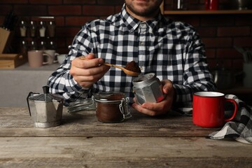 Man putting ground coffee into moka pot at wooden table in kitchen, closeup