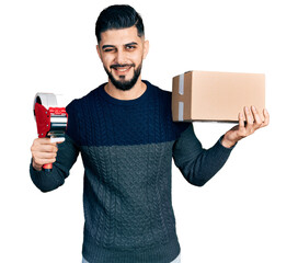 Young arab man with beard holding packing tape and cardboard box winking looking at the camera with...