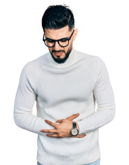 Young arab man with beard wearing elegant turtleneck sweater and glasses with hand on stomach...