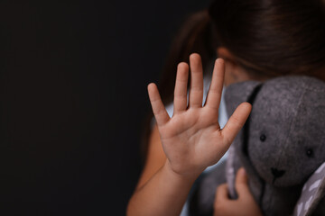 Child abuse. Little girl with toy bunny doing stop gesture on dark background, selective focus. Space for text