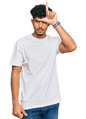 Young arab man wearing casual white t shirt making fun of people with fingers on forehead doing loser gesture mocking and insulting.