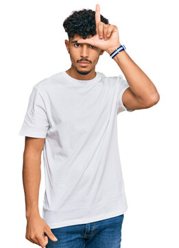 Young arab man wearing casual white t shirt making fun of people with fingers on forehead doing loser gesture mocking and insulting.