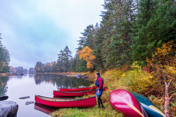 young man with canoes on the shore of a calm reflective part of the lower madawaska river ontario in fall with autumn color room for text - obrazy, fototapety, plakaty