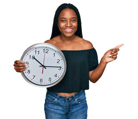 Young african american woman holding big clock smiling happy pointing with hand and finger to the...