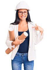 Young beautiful latin girl wearing architect hardhat holding blueprints pointing finger to one self smiling happy and proud