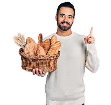 Young hispanic man with beard holding wicker basket with bread smiling happy pointing with hand and finger to the side