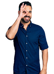 Young hispanic man with beard wearing casual blue shirt smiling happy doing ok sign with hand on eye looking through fingers