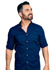 Young hispanic man with beard wearing casual blue shirt looking away to side with smile on face, natural expression. laughing confident.