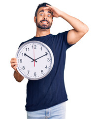 Young hispanic man holding big clock stressed and frustrated with hand on head, surprised and angry...