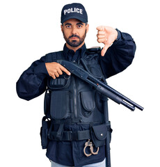 Young hispanic man wearing police uniform holding shotgun with angry face, negative sign showing dislike with thumbs down, rejection concept