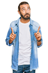 Attractive man with long hair and beard wearing casual denim jacket pointing up looking sad and...