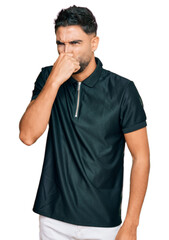 Young man with beard wearing sportswear smelling something stinky and disgusting, intolerable smell, holding breath with fingers on nose. bad smell