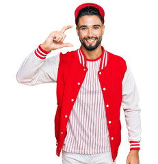 Young man with beard wearing baseball uniform smiling and confident gesturing with hand doing small size sign with fingers looking and the camera. measure concept.