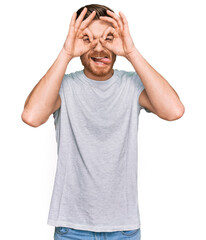 Young redhead man wearing casual grey t shirt doing ok gesture like binoculars sticking tongue out, eyes looking through fingers. crazy expression.