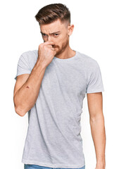 Young redhead man wearing casual grey t shirt smelling something stinky and disgusting, intolerable smell, holding breath with fingers on nose. bad smell