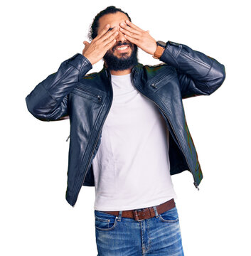 Young arab man wearing casual leather jacket covering eyes with hands smiling cheerful and funny. blind concept.