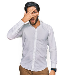 Young hispanic man wearing business shirt and glasses peeking in shock covering face and eyes with hand, looking through fingers with embarrassed expression.