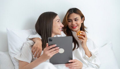 Young Asian lesbian couple sitting on bed during happy resting time, using tablet, exploring social...