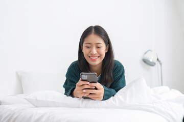 Cute Asian woman lying on bed using phone, exploring social media in the morning.