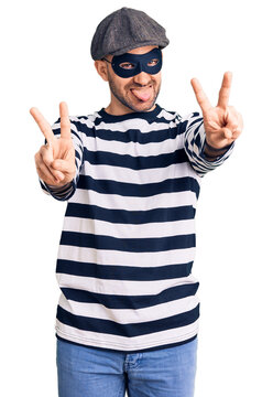 Young handsome man wearing burglar mask smiling with tongue out showing fingers of both hands doing victory sign. number two.
