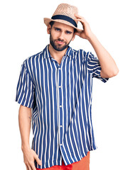 Young handsome man with beard wearing summer hat and striped shirt confuse and wonder about question. uncertain with doubt, thinking with hand on head. pensive concept.