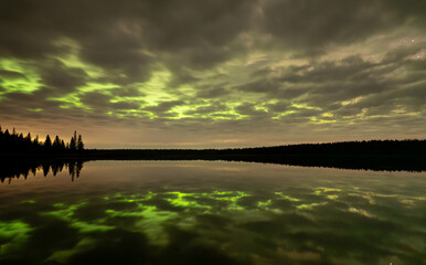 A night sky with heavy clouds that are backlit by bright green Aurora. The clouds and Aurora are...