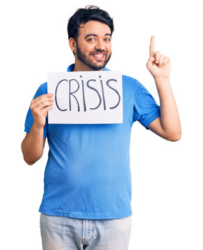 Young hispanic man holding crisis message paper surprised with an idea or question pointing finger with happy face, number one
