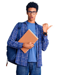 Young african american man wearing student backpack holding book surprised pointing with hand finger to the side, open mouth amazed expression.