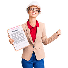Young beautiful woman wearing architect hardhat holding clipboard with contract document screaming proud, celebrating victory and success very excited with raised arms