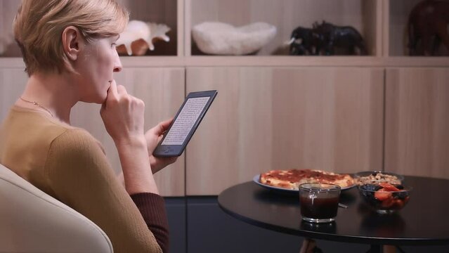An adult woman excitedly reads an e-book at the table at home, forgetting about coffee