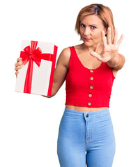 Young blonde woman holding gift with open hand doing stop sign with serious and confident expression, defense gesture